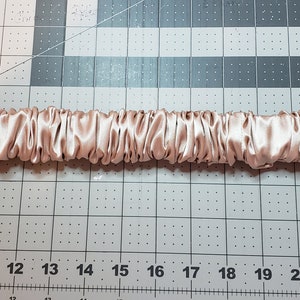 May include: A close-up of a long, narrow strip of pale pink satin fabric, gathered into a series of ruffles. The fabric is laid out flat on a gridded surface.