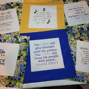 May include: A collection of six fabric squares with bible verses embroidered on them. The squares are arranged in a grid pattern on a table. The verses are: "She is far more precious than rubies", "The LORD will give strength unto his people; The LORD will bless his people with peace. Psalm 29:11", "I will sing of mercy and judgement. Unto thee, O LORD, will I sing. Psalm 101:1", "In the beginning was the Word, and the Word was with God, and the Word was God. John 1:1", "They that wait upon the LORD shall renew their strength; they shall mount up with wings of eagles; they shall run and not be weary; they shall walk and not be faint. Isaiah 40:31", and "Her children arise up, and call her blessed: Her husband also, and he praiseth her. Many daughters have done virtuously, but thou excellest them all. Proverbs 31:28-29".