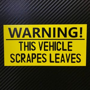 May include: A yellow warning sign with black text that reads "WARNING! THIS VEHICLE SCRAPES LEAVES".