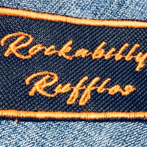 Peut inclure: Un écusson brodé noir et orange avec le texte "Rockabilly Ruffians" en écriture cursive.