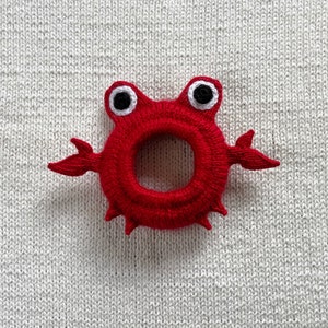May include: Red knitted crab toy with white eyes and a ring shape in the center.