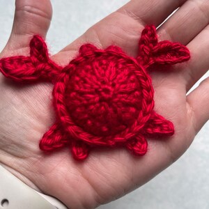 May include: A handmade, bright red crochet crab. The crab has a circular body with eight legs and two pincers. The crab is made of a soft, textured yarn. The crab is a decorative item.