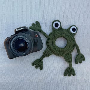 May include: A black Canon EOS Rebel T3i digital camera with an 18-55mm lens positioned next to a dark green knitted frog-shaped hair scrunchie featuring two white and black eyes and four splayed limbs with webbed-style fingers and toes.