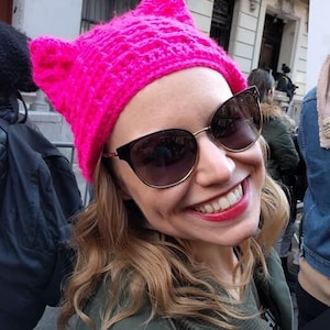 May include: A woman wearing a bright pink, cat-ear knit hat and sunglasses smiles at the camera. The hat is a vibrant fuchsia color. The sunglasses have a black frame with gold accents. She is wearing a green jacket.