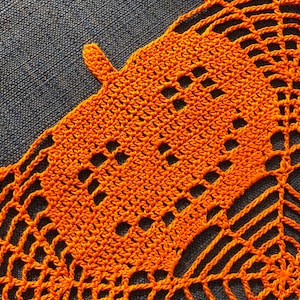 May include: Close-up of an orange crocheted doily with a detailed openwork design. The handmade piece features a repeating pattern of squares and curved lines, creating a delicate, lacy texture. The doily is made of a vibrant orange yarn.