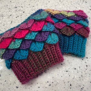 May include: A pair of colorful crocheted wrist warmers with a scale pattern. The warmers are made with a variety of shades of blue, purple, pink, and green yarn.