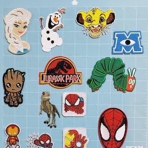 May include: A collection of embroidered patches featuring various cartoon characters and logos. Includes Elsa, Olaf, Simba, Groot, a dinosaur, Spider-Man, Iron Man, and the Jurassic Park logo. The patches are colorful and detailed, suitable for clothing or accessories.