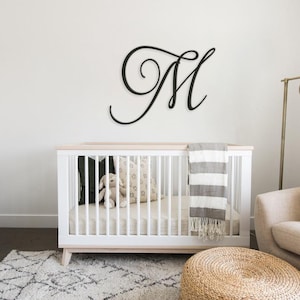 May include: A white crib with a gray and white striped blanket and a stuffed animal in the crib. A black metal letter M is on the wall above the crib. There is a white dresser to the left of the crib and a beige armchair to the right.
