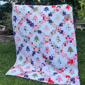 May include: A quilted blanket featuring a white background with a repeating pattern of colorful Christmas trees in shades of pink, green, and blue. The design includes small printed motifs of Santa Claus and holiday scenes within the tree shapes.
