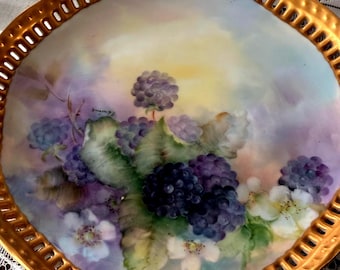 Peut inclure: Une assiette décorative à bordure dorée avec un motif peint à la main. Le motif présente des grappes de mûres violet foncé, des feuilles vertes et des fleurs blanches sur un fond pastel. Le bord de l'assiette a un motif ajouré.