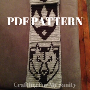May include: A crocheted scarf featuring three different designs. The top design is a white horse head on a brown background. The middle design is a white triangle with a brown centre. The bottom design is a white wolf head on a brown background. The text "PDF PATTERN" is printed on the scarf.