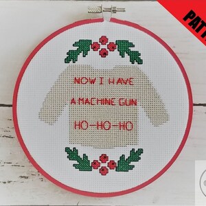 Könnte beinhalten: Ein weißes Kreuzstichmuster in einem roten Stickrahmen mit einem beigefarbenen Pullover-Design, auf dem "NOW I HAVE A MACHINE GUN HO-HO-HO" steht. Der Pullover ist von grünen Stechpalmenblättern und roten Beeren umgeben.