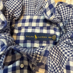 May include: A blue and white gingham button-down shirt with a ruffled collar. The interior neck label features the Ralph Lauren logo and text, with a size M/M label indicating it was made in the Philippines. A single white button is visible.