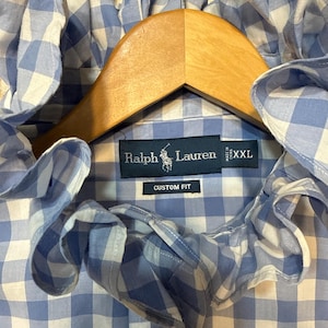 May include: A light blue and white gingham button-down shirt featuring a ruffled collar. The interior label displays the text Ralph Lauren, Custom Fit, and XXL. The shirt is hanging on a wooden hanger, showcasing the checkered pattern clearly.