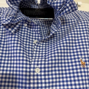 May include: A blue and white gingham button-down shirt with a ruffled collar. The shirt has a classic check pattern and a small embroidered logo of a polo player on a horse. The shirt is buttoned up and hanging on a black hanger.