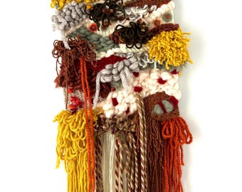 May include: A textured wall hanging with a wooden dowel at the top. The woven artwork features a variety of colors, including yellow, orange, brown, gray, and white. The design incorporates knotted and fringed elements, creating a tactile and visually interesting piece.