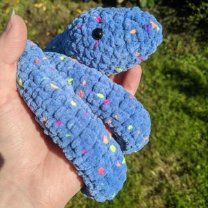 May include: A blue crochet snake plush toy made from soft, textured yarn. The snake features a speckled pattern with small, vibrant flecks of pink, orange, yellow, and green throughout the blue material. It has a single visible black plastic eye.