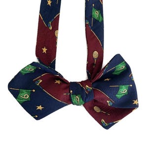 Può includere: Un papillon blu navy e bordeaux a tema golf. La cravatta presenta un motivo ripetuto di bandiere da golf con il numero 9, mazze da golf e stelle dorate. Il papillon è realizzato in materiale setoso.