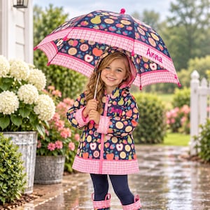 May include: A navy blue hooded raincoat, matching rain boots, and an umbrella, all featuring a colorful fruit pattern of apples, pears, and strawberries. The raincoat and umbrella are accented with pink plaid trim and the name Anna in pink text.