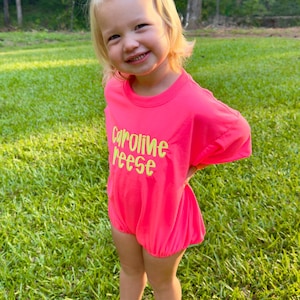 May include: A child in a bright pink romper with the name "Caroline Reese" in neon green. The child wears white sandals and a large white bow. The background features a grassy lawn and trees, typical of a park or garden setting.