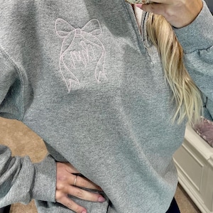May include: A gray sweatshirt with a quarter-zip neckline and a light pink embroidered bow design. The sweatshirt has long sleeves and a relaxed fit. The zipper is silver. The overall style is casual and comfortable.
