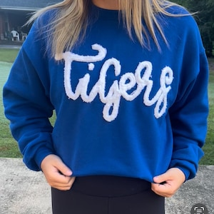 May include: A royal blue sweatshirt with the word "Tigers" in white, fluffy, cursive lettering across the chest. The sweatshirt is a solid color and has a crew neck. The person is wearing black leggings.