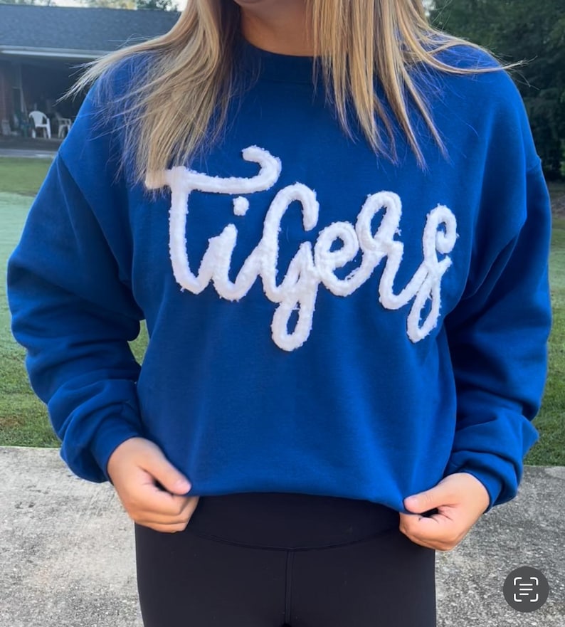 May include: A royal blue sweatshirt with the word "Tigers" in white, fluffy, cursive lettering across the chest. The sweatshirt is a solid color and has a crew neck. The person is wearing black leggings.