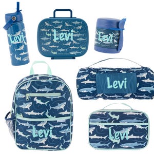 May include: A blue shark-themed lunch bag set with a backpack, lunch bag, water bottle, thermos, and a nap mat. The set is personalized with the name "Levi".