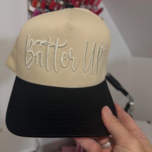 May include: A tan and black baseball cap with the embroidered text "Batter Up" in a cursive font. The cap has a flat brim and a structured design. The hat is being held by a person.