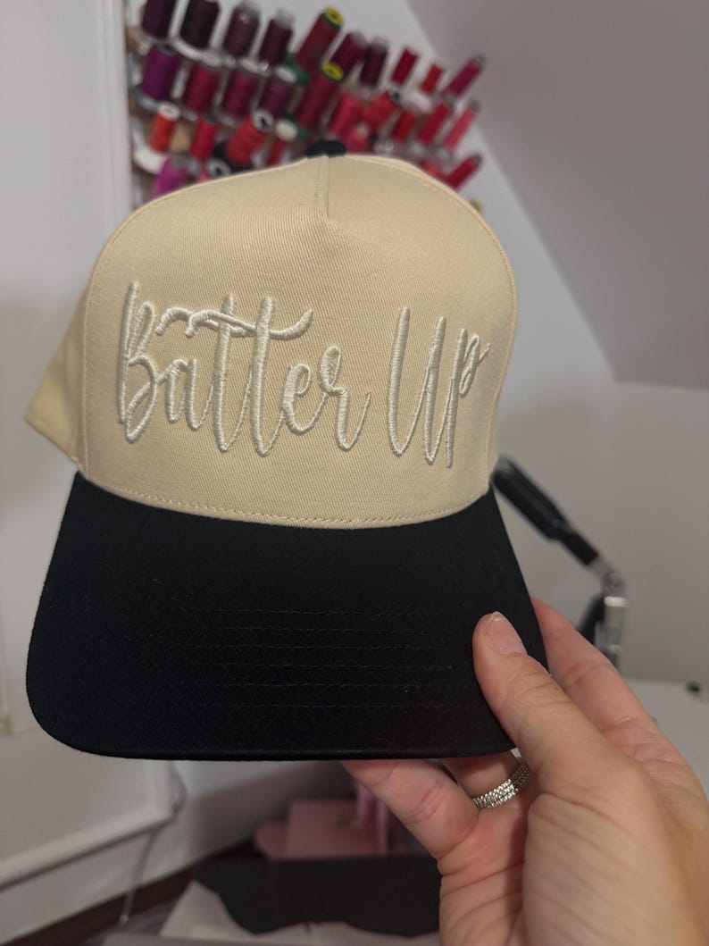 May include: A tan and black baseball cap with the embroidered text "Batter Up" in a cursive font. The cap has a flat brim and a structured design. The hat is being held by a person.