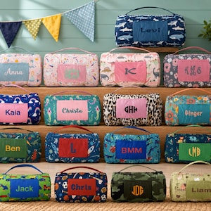 May include: A collection of personalized blankets with various patterns and colors. Each blanket features a rectangular patch with a name or initials in a contrasting color. Designs include rainbows, dinosaurs, and floral prints. The blankets have a handle for easy carrying.