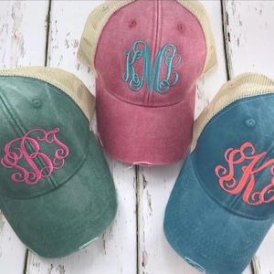 May include: Three distressed baseball caps with mesh back panels. The caps are in different colors: teal, pink, and blue. Each cap has a different monogram embroidered on the front in a contrasting color.