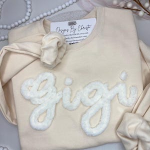 May include: A cream-coloured sweatshirt with the word "Gigi" in white fluffy lettering. The sweatshirt has a small tag on the front that says "DBC Designs By Christa" with a website address and social media handles.