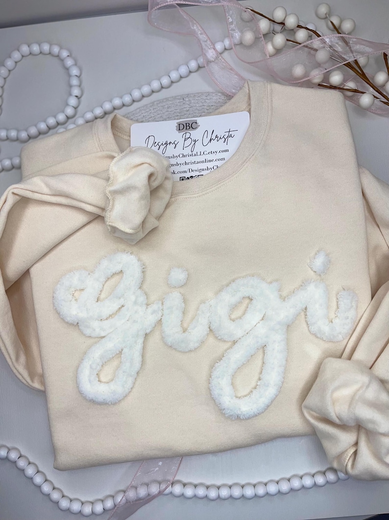 May include: A cream-coloured sweatshirt with the word "Gigi" in white fluffy lettering. The sweatshirt has a small tag on the front that says "DBC Designs By Christa" with a website address and social media handles.