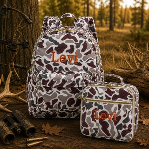 May include: A backpack and a matching lunch bag, both featuring a camouflage pattern in shades of white, gray, and dark brown. Each item is embroidered with the name Levi in orange thread. The bags have gold-toned zippers and top handles.