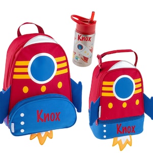 May include: A red, blue, and yellow rocket-themed backpack, lunch bag, and water bottle set. The backpack and lunch bag feature a large circular window and the name "Knox". The water bottle has a red lid and a rocket graphic.