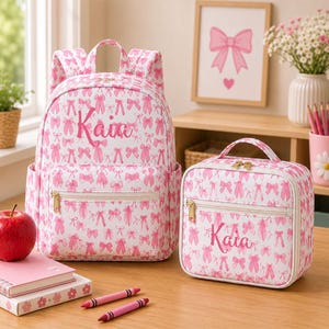 Könnte beinhalten: Ein rosa-weißes Rucksack- und Lunchpaket-Set. Der Rucksack und die Lunchtasche sind mit einem Muster aus rosa Schleifen und Ballettschuhen bedeckt. Der Name "Kaia" ist in rosa Schrift auf beiden Artikeln aufgedruckt. Beide Artikel haben goldene Reißverschlüsse.