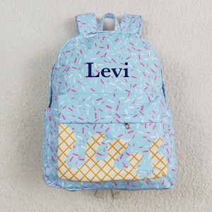 May include: A light blue backpack with a sprinkle pattern and the name "Levi" in navy blue. The front pocket features a waffle cone design with a gold grid pattern and white and pink accents. Includes two shoulder straps and a top handle.