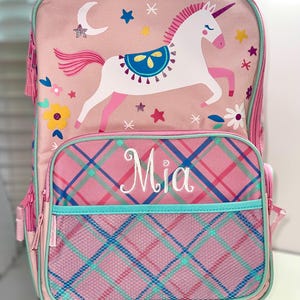 May include: Pink rolling backpack with a unicorn design. The backpack features a white unicorn with a pink mane and tail, surrounded by stars and flowers. The front pocket has the name "Mia" embroidered on it.