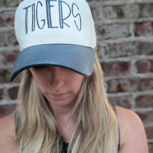 Op de afbeelding: Een witte en grijze baseballcap met het woord "TIGERS" in blauw geborduurd. De cap heeft een gebogen klep en een gestructureerde kroon. De achtergrond is een bakstenen muur.