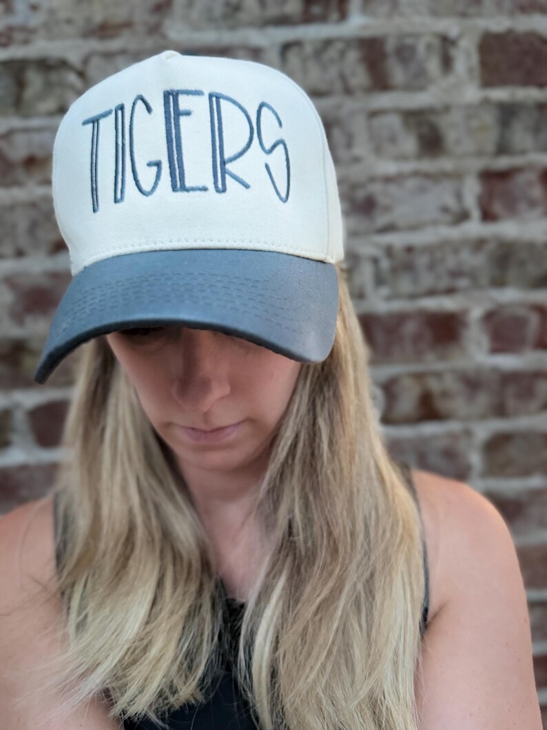 Op de afbeelding: Een witte en grijze baseballcap met het woord "TIGERS" in blauw geborduurd. De cap heeft een gebogen klep en een gestructureerde kroon. De achtergrond is een bakstenen muur.