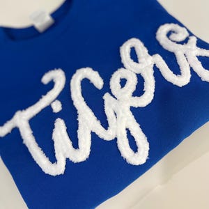 May include: Royal blue sweatshirt with the word "Tigers" in white, textured, cursive lettering. The sweatshirt is folded neatly on a white surface. The text is the focal point, creating a bold contrast against the solid blue background.