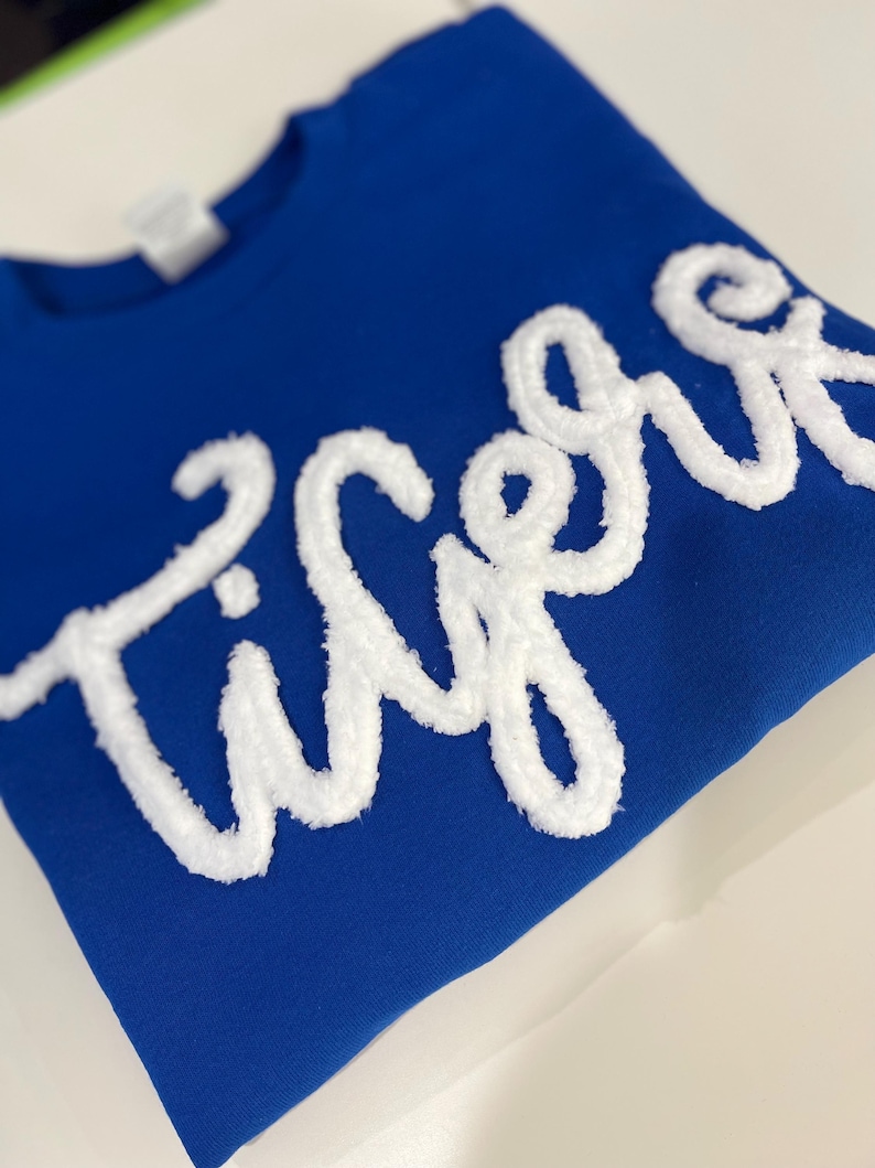 May include: Royal blue sweatshirt with the word "Tigers" in white, textured, cursive lettering. The sweatshirt is folded neatly on a white surface. The text is the focal point, creating a bold contrast against the solid blue background.