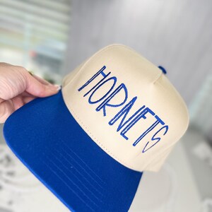 Op de afbeelding: Een beige en blauwe baseballcap met het woord "HORNETS" in blauw geborduurd. De cap heeft een blauwe klep en een beige kroon. De cap is een sportthema accessoire.