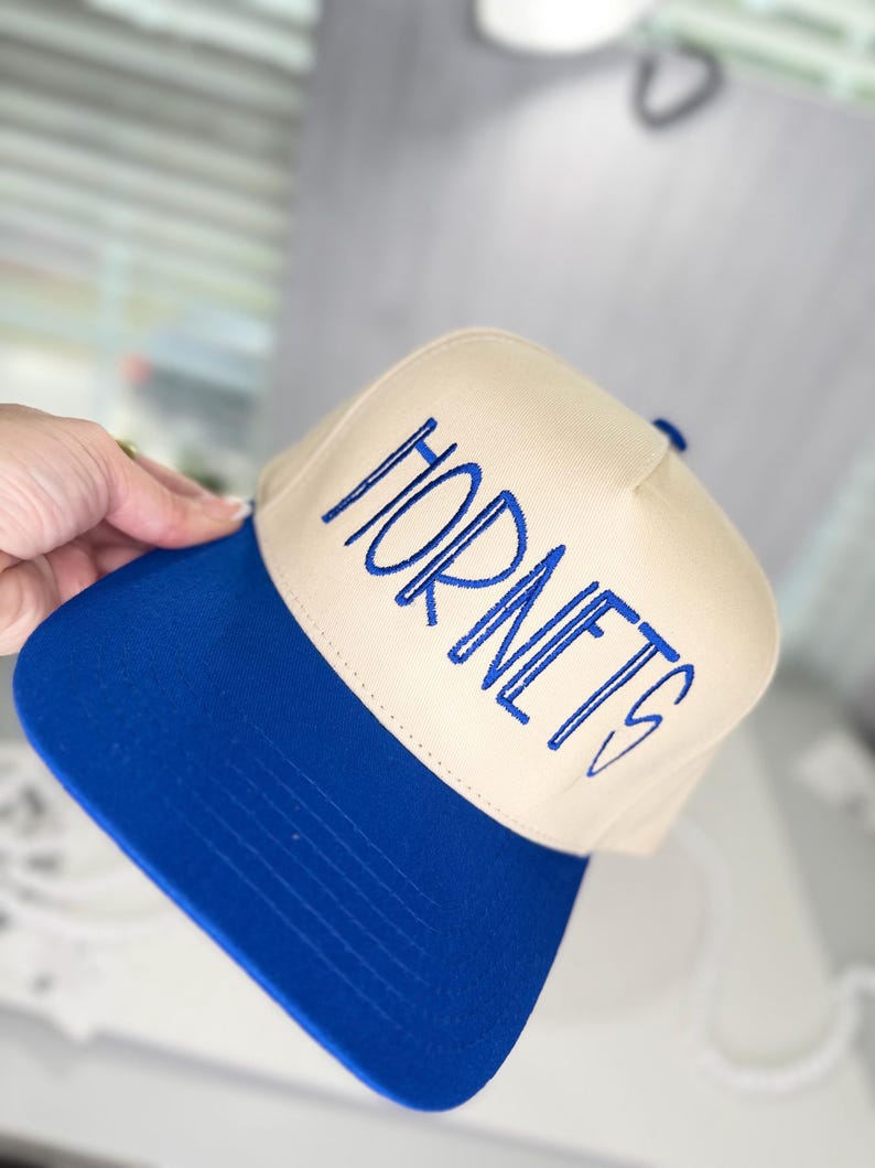 Op de afbeelding: Een beige en blauwe baseballcap met het woord "HORNETS" in blauw geborduurd. De cap heeft een blauwe klep en een beige kroon. De cap is een sportthema accessoire.