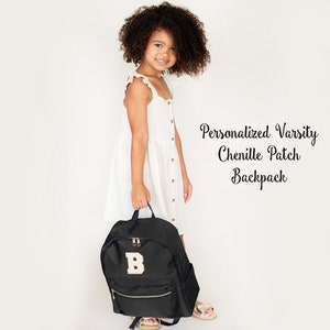 May include: Black backpack with a gold zipper and a chenille patch letter "B". The backpack has a front pocket and a top handle. The text "Personalized Varsity Chenille Patch Backpack" is displayed in the image.