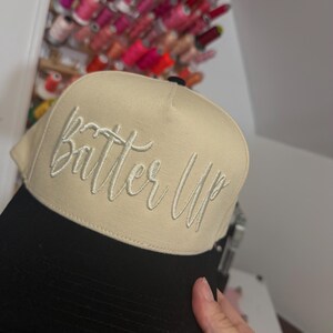 May include: A beige and black baseball cap with the embroidered text "Batter Up" in a cursive font. The cap has a curved brim and a structured crown. The background shows spools of thread.