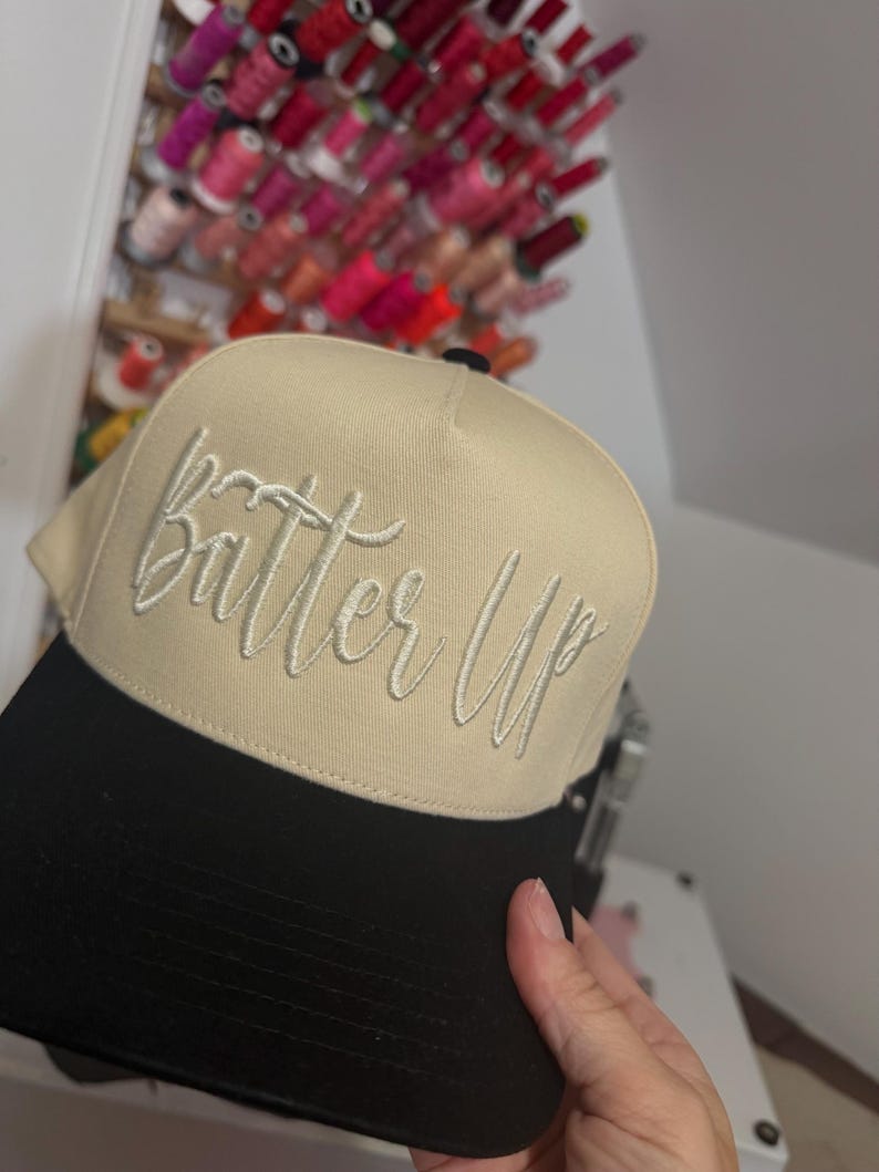 May include: A beige and black baseball cap with the embroidered text "Batter Up" in a cursive font. The cap has a curved brim and a structured crown. The background shows spools of thread.