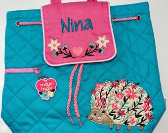 Puede incluir: Una mochila acolchada de color azul turquesa con un cordón de ajuste rosa y una solapa rosa con el nombre "Nina" bordado. La mochila tiene un erizo con flores bordadas y un dije de corazón rosa que dice "hedge hugs!" colgando de la cremallera.