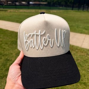 May include: A beige and black baseball cap with the text "Batter Up" embroidered in white script. The cap has a structured front panel and a curved brim. The cap is being held up in front of a green field.